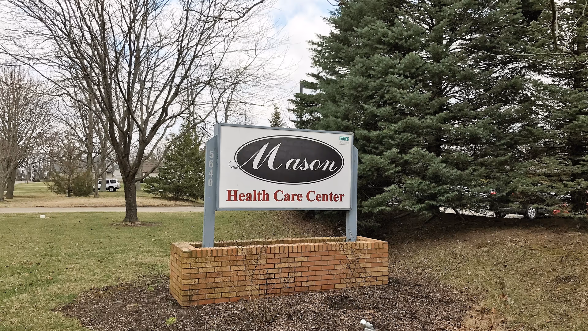 Outdoor view of a sign for Mason Health Care Center mounted on a brick base with trees and grass surrounding it.