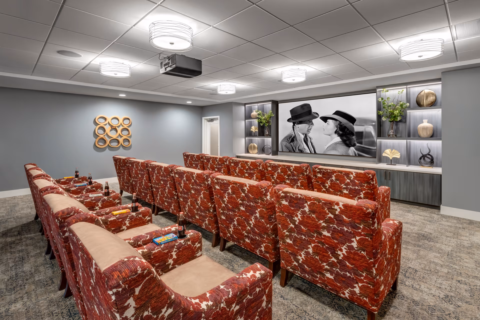 A cozy home theater room with two rows of red patterned armchairs facing a large screen showing a black and white movie scene of a man and woman in hats. The room has gray walls, decorative shelves with vases and plants on either side of the screen, and modern ceiling lights.