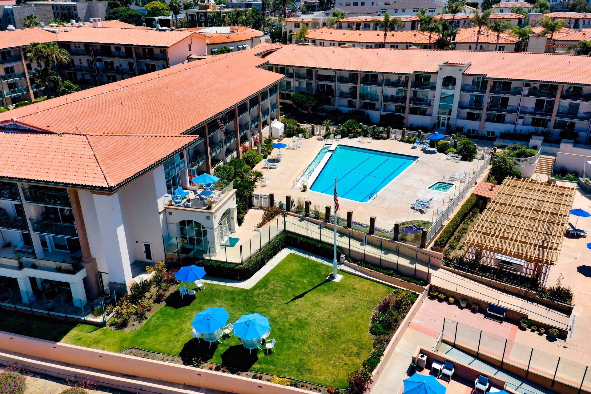 Aerial view of White Sands La Jolla senior living facility showing a large building with red tile roofs surrounding a courtyard area. The courtyard features a swimming pool, a hot tub, several blue umbrellas with tables and chairs, a green lawn with more blue umbrellas, and a pergola-covered seating area. Palm trees and other greenery are visible around the property.