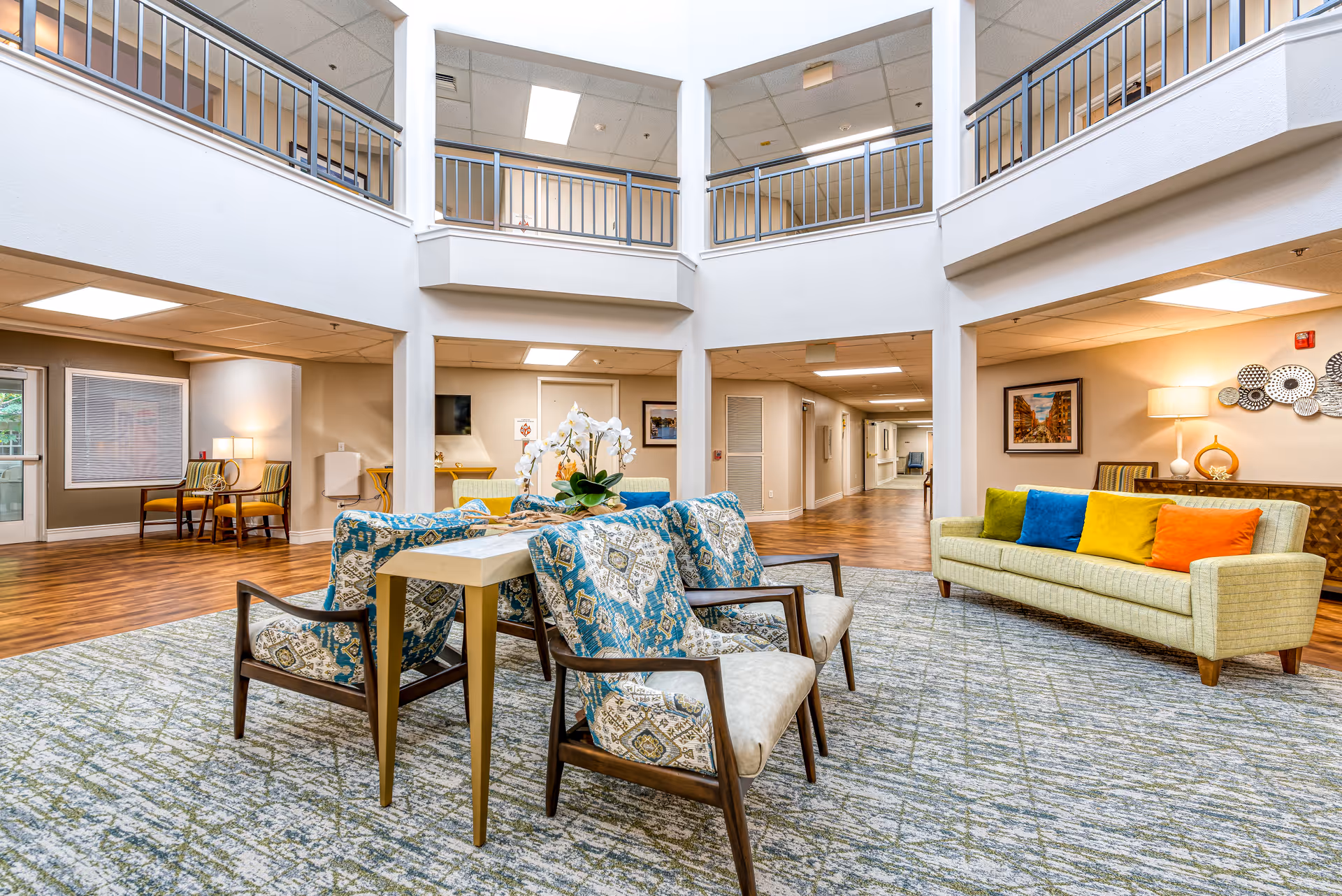 A bright and spacious senior living facility common area with a high ceiling and an open second-floor balcony. The room features a seating arrangement with four patterned armchairs around a small table with a white orchid centerpiece, and a light green sofa adorned with colorful cushions in blue, yellow, and orange. The floor is covered with a patterned carpet, and the walls are decorated with framed artwork and a lamp on a side table. The area is well-lit with ceiling lights and natural light from windows.