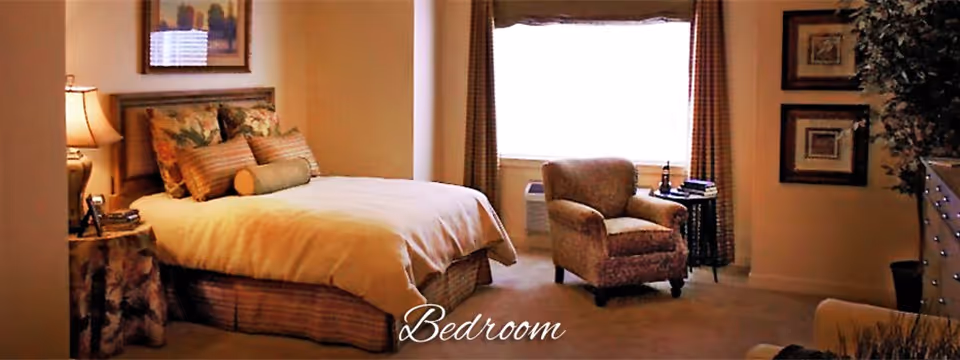 Furnished bedroom with a bed, bedside table and lamp, an armchair by a window, and decorative wall art.