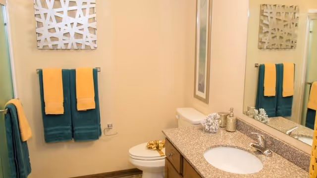 A clean bathroom with a granite countertop sink, a toilet, and a large mirror. The walls are beige, and there are teal and yellow towels hanging on towel racks. A decorative white wall art piece is above the towels.