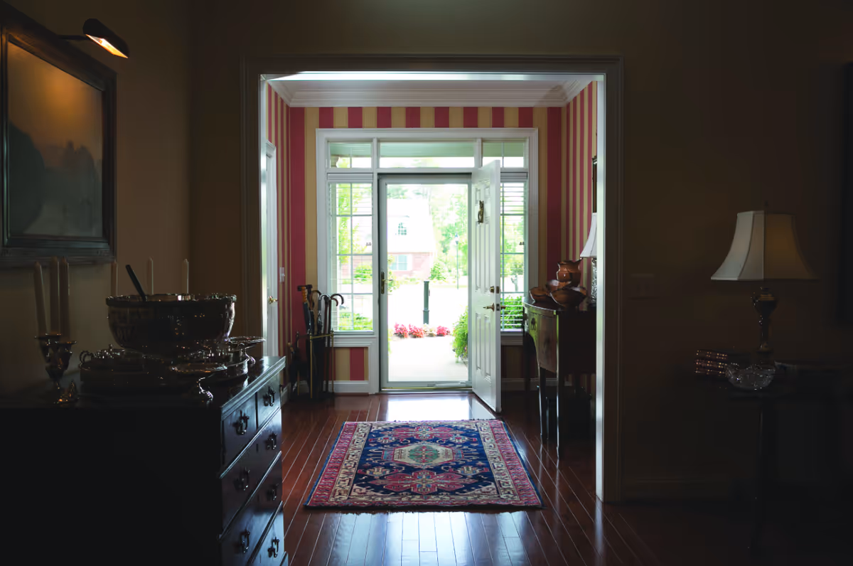 A foyer with a patterned rug on hardwood floors leading to an open front door with striped wallpaper and side furniture and lamps.