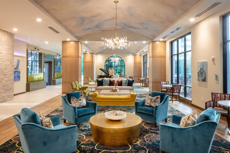 A spacious and elegant living room area with a high ceiling painted with a sky mural. The room features a large chandelier, teal armchairs with patterned pillows, a yellow sofa, and a round gold coffee table with a decorative bowl. Large windows on the right side allow natural light to fill the room, and there is a grand piano in the background near an arched window. Additional seating areas with chairs and tables are visible along the sides.