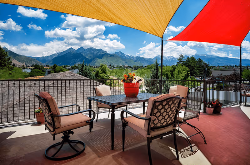 Outdoor patio area with a table and four cushioned chairs under red and yellow shade sails, overlooking a scenic view of green trees and mountains under a blue sky with scattered clouds.
