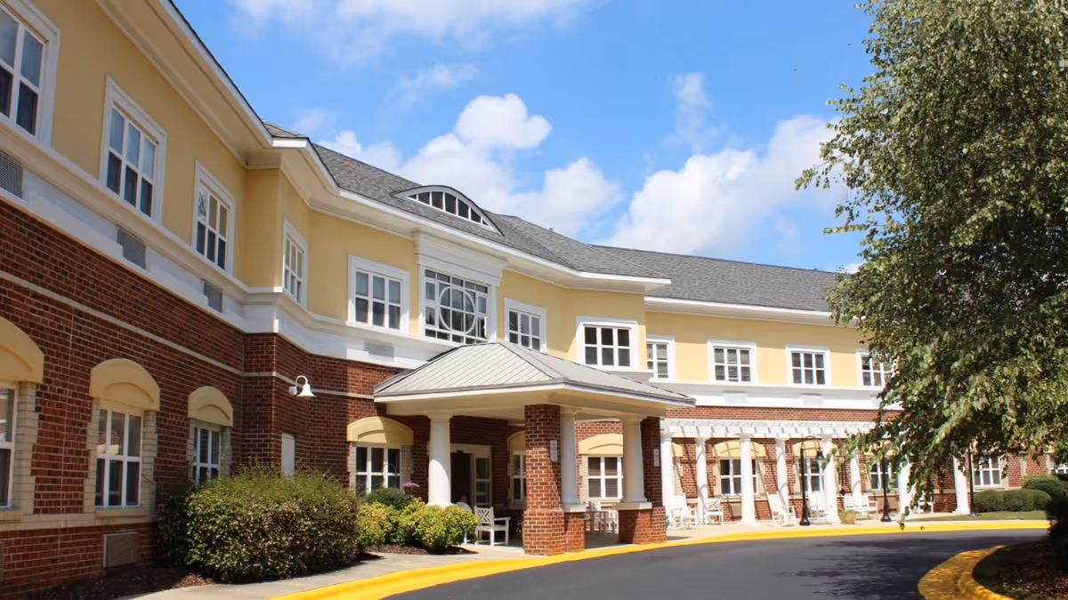 Exterior view of a senior living community building with a brick and yellow facade, multiple windows, a covered entrance supported by columns, bushes, and a tree on the right side under a partly cloudy blue sky.