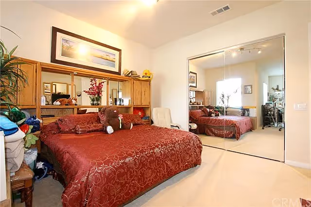 A bedroom with a large bed covered in a red patterned bedspread, a stuffed bear on the bed, wooden headboard with shelves, a framed picture above the bed, a white chair beside the bed, and mirrored sliding closet doors reflecting the room.