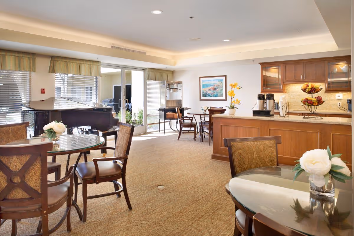 A bright and inviting common area with several round glass-top tables and wooden chairs, a grand piano near large windows with blinds, a counter with coffee dispensers and a fruit basket, and a framed picture on the wall. The room has beige carpeting and soft lighting.