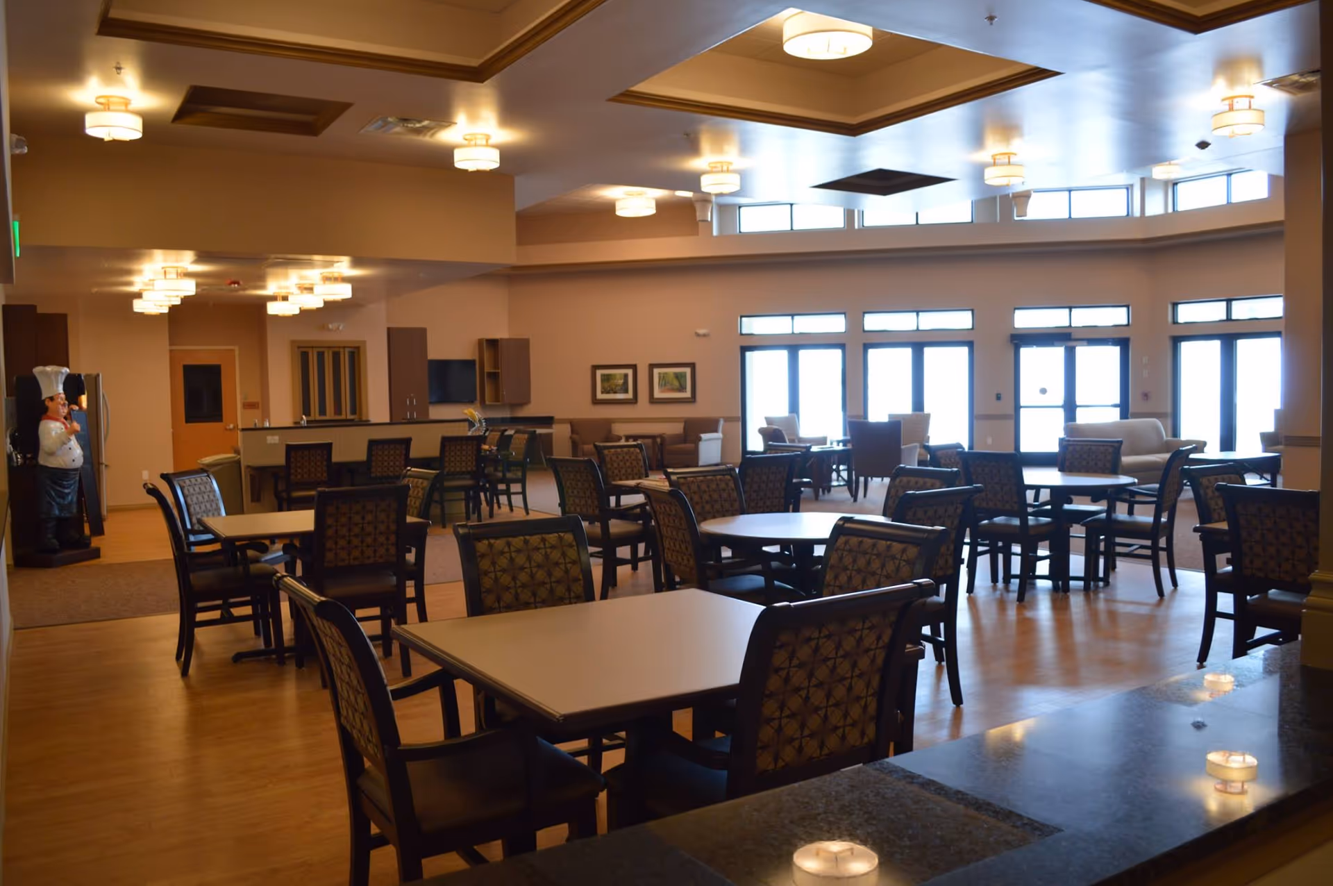 Spacious dining and common area with multiple tables and chairs, large windows, and warm overhead lighting.