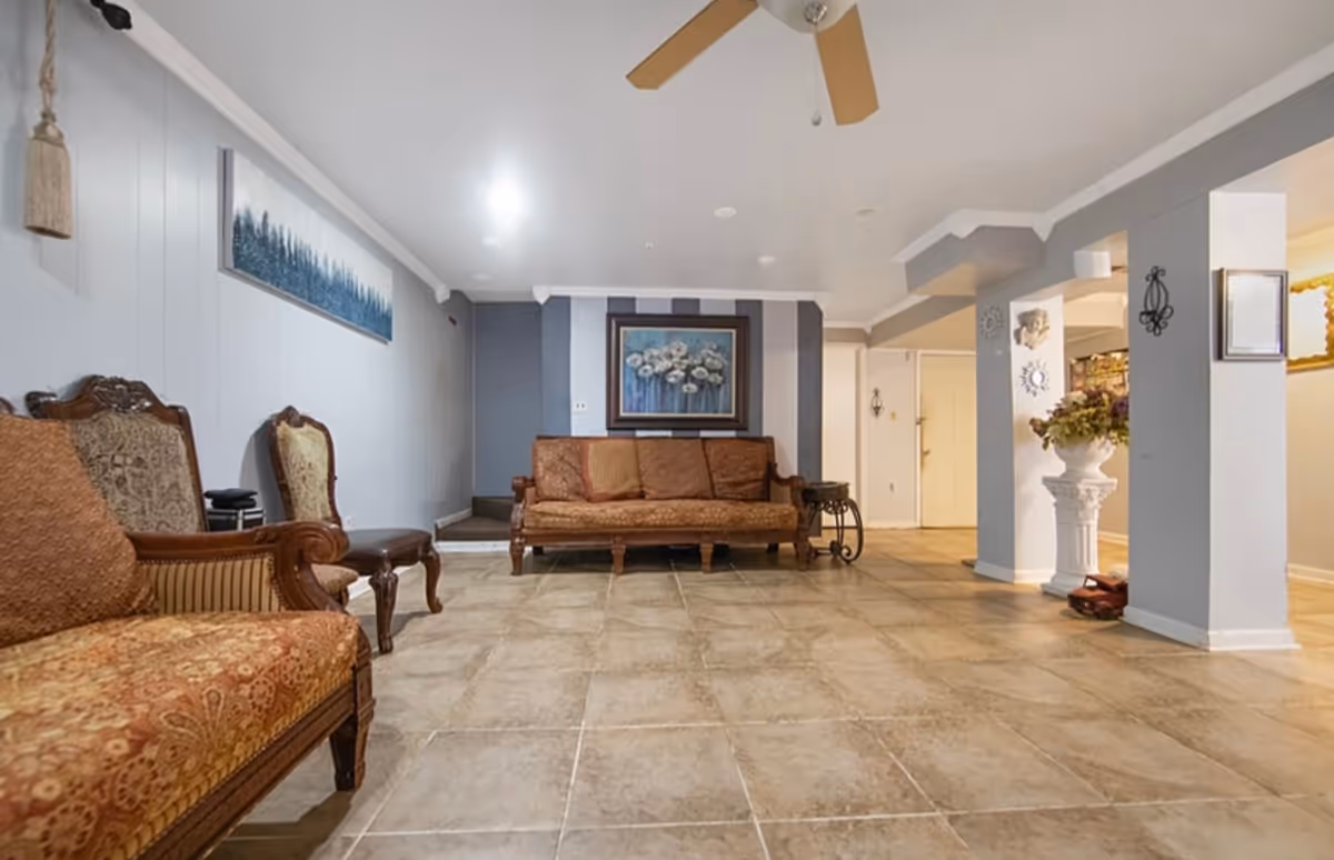 A spacious living room with tiled flooring, featuring vintage-style upholstered chairs and a matching sofa. The walls are painted in light gray and white with decorative paintings and wall hangings. A ceiling fan with wooden blades is mounted on the white ceiling. There is a decorative white pedestal with a floral arrangement near a column on the right side.