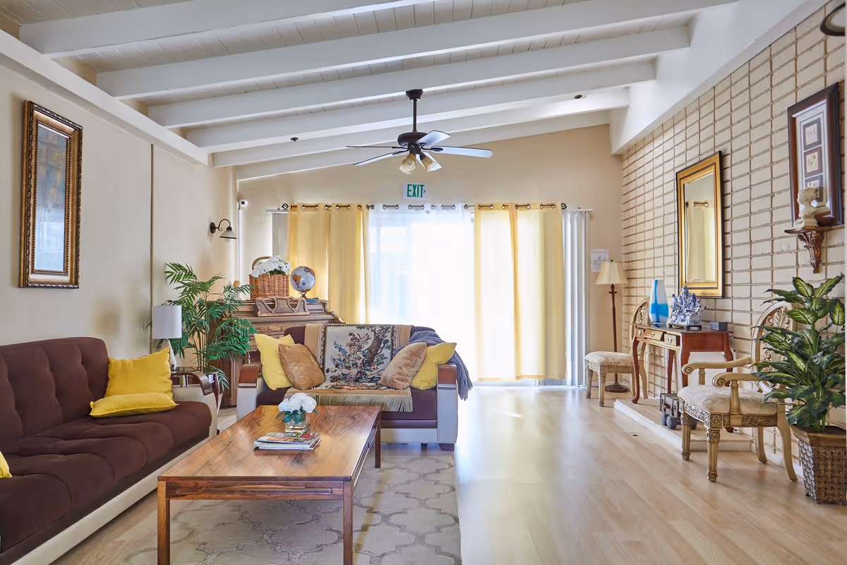 A bright and cozy living room with a brown sofa and loveseat adorned with yellow and beige cushions. A wooden coffee table with a small flower arrangement and books sits on a patterned rug. The room features light wood flooring, a ceiling fan, potted plants, framed artwork on the walls, and large windows covered with sheer and yellow curtains letting in natural light.