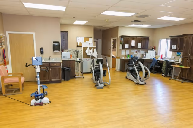 A rehabilitation room with exercise equipment including stationary bikes and a pedal exerciser. The room has wooden flooring, beige walls, and wooden cabinets along the back wall. There are desks with office chairs and various papers and supplies on the counters. A bulletin board with pinned papers is visible on the wall near a closed wooden door.