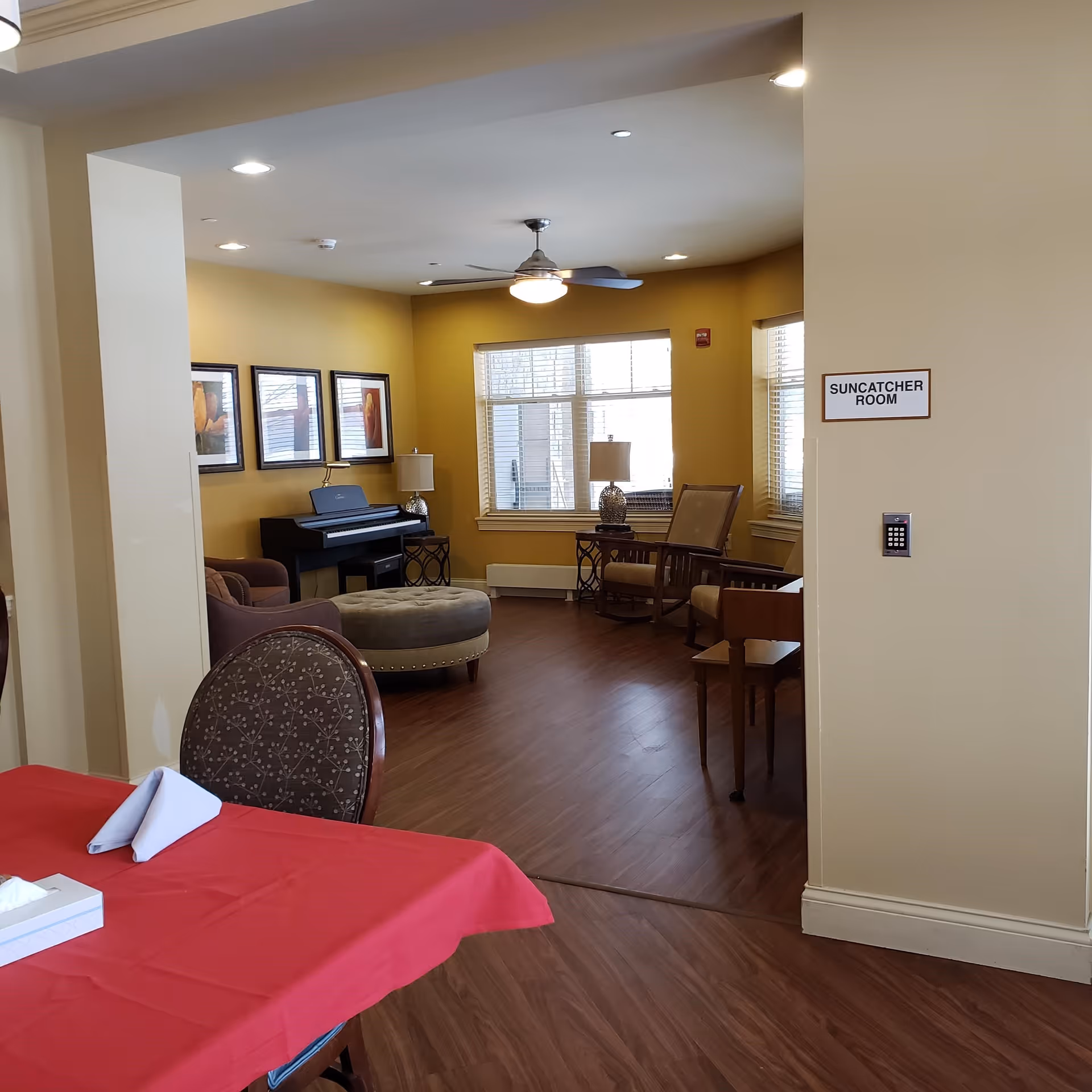 Interior view of a senior living facility room named 'Suncatcher Room' with wooden flooring, beige walls, a ceiling fan with light, a piano against the wall, framed artwork, a round ottoman, chairs, side tables with lamps, and large windows letting in natural light. In the foreground, there is a table with a red tablecloth and a chair.