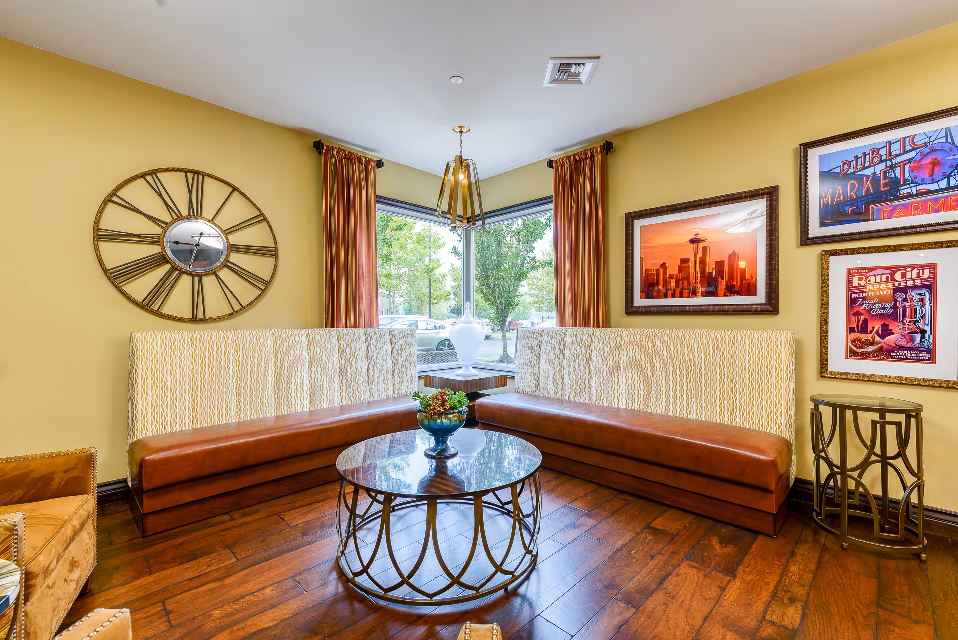 A cozy seating area with two brown and patterned cushioned benches arranged in an L-shape around a round glass coffee table with a decorative plant. The walls are painted yellow and decorated with a large round clock and framed pictures. There is a large corner window with red and yellow striped curtains letting in natural light.