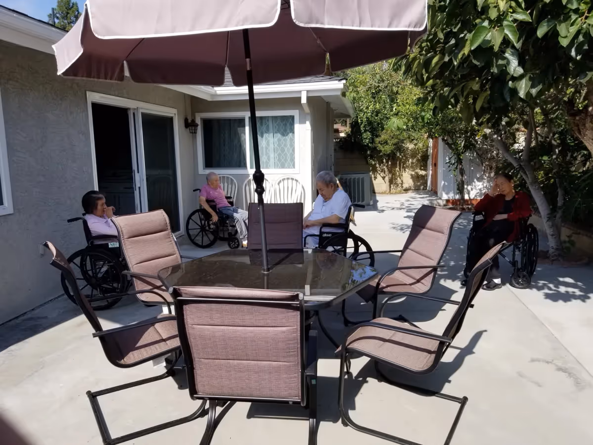 Outdoor patio area at a senior living facility with four elderly individuals in wheelchairs sitting around the perimeter. There is a glass-top table with six chairs and a large umbrella providing shade. The area is paved with concrete and surrounded by a building wall and greenery.