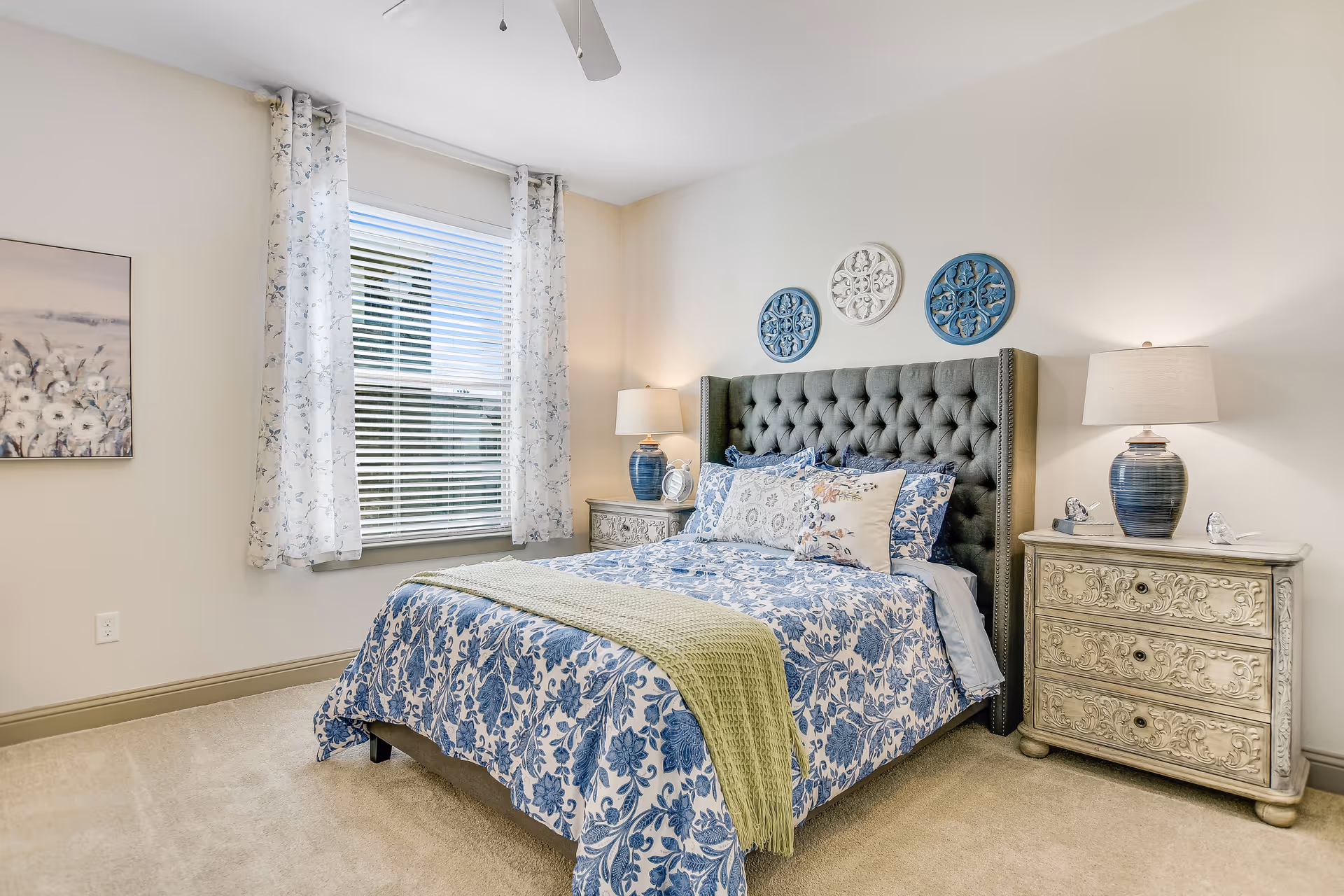 A bedroom with a large tufted headboard bed dressed in blue and white floral bedding and multiple pillows. There are two ornate nightstands on either side of the bed, each with a blue lamp. A window with white blinds and floral curtains is on the left wall, and three decorative round wall hangings are above the headboard. The room has beige walls and carpeted flooring.