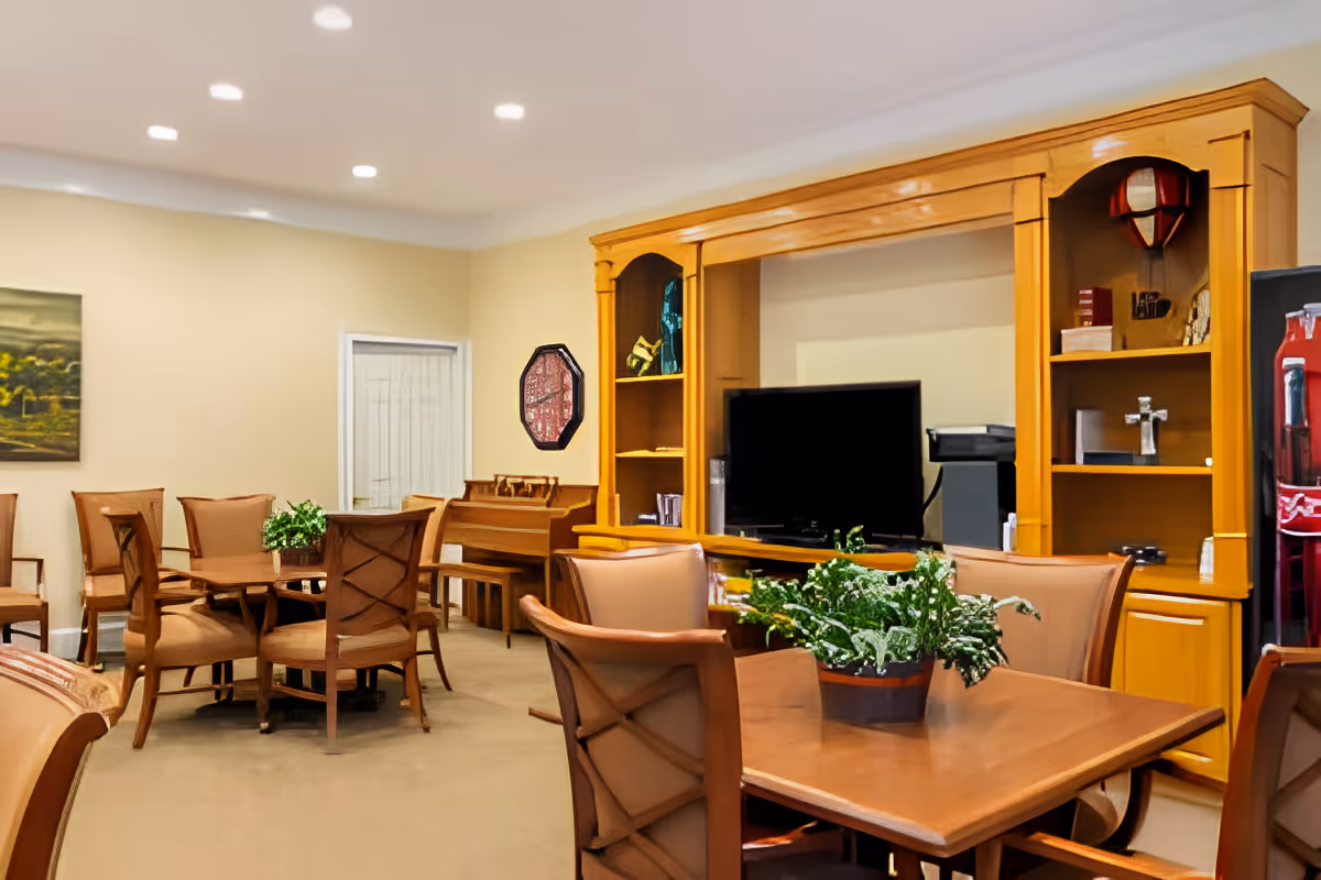 A cozy senior living common area with wooden tables and chairs, a large wooden entertainment center with a television, shelves with decorative items, a piano in the corner, and potted plants on the tables. The room has beige walls and carpeted floors with recessed lighting in the ceiling.