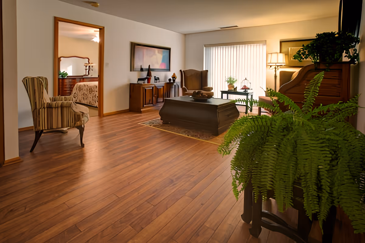 Spacious living room with wood floors, seating, a coffee table, and large potted ferns with a bedroom visible through an open doorway.