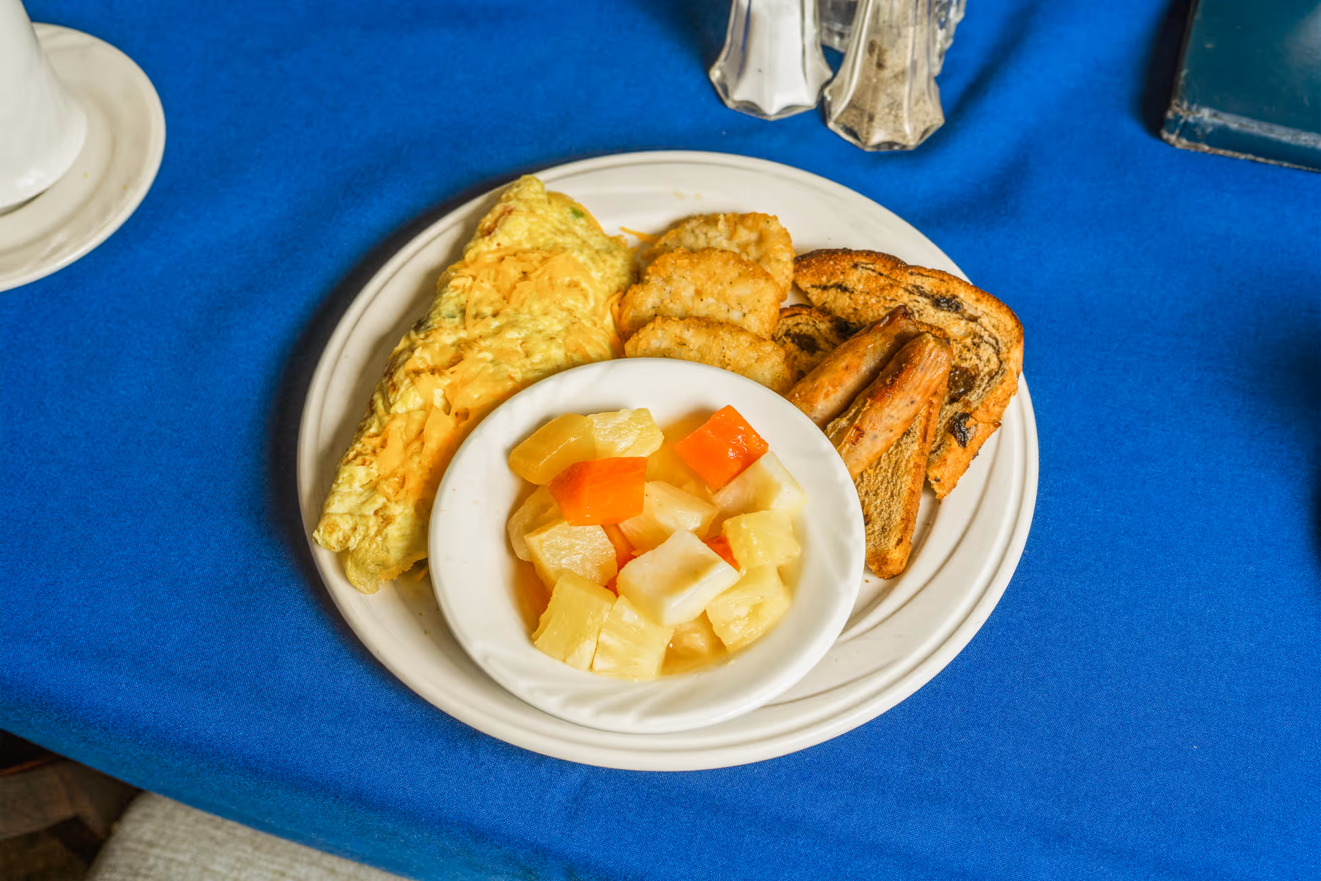 Plate with an omelette, hash browns, toast, sausages, and a bowl of mixed fruit on a blue tablecloth.