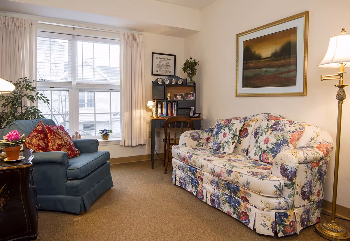Cozy living room with a floral-patterned sofa, blue armchair, window, small desk and floor lamp.