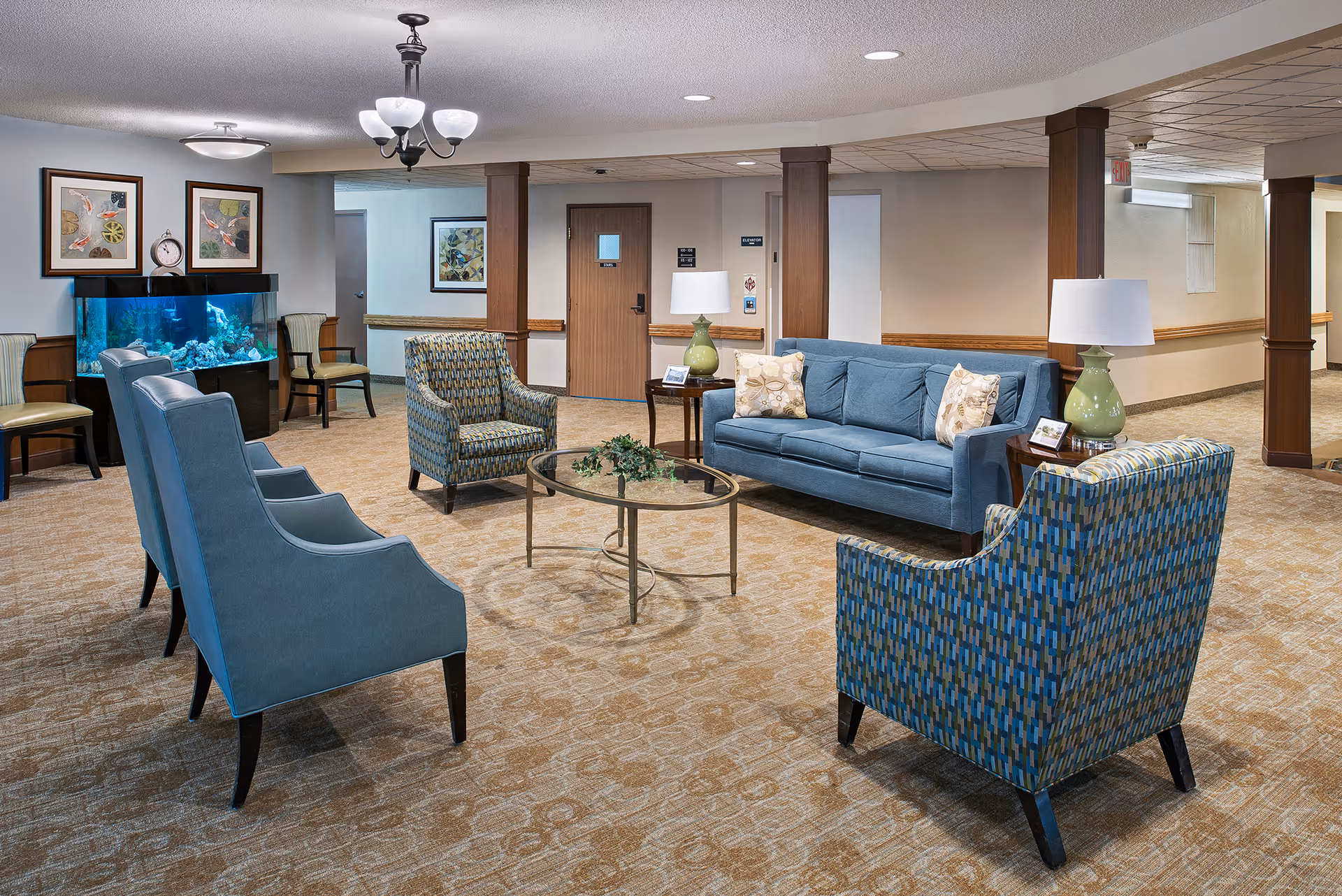 Spacious assisted-living lobby with blue upholstered sofa and chairs arranged around a glass coffee table and an aquarium along the back wall.