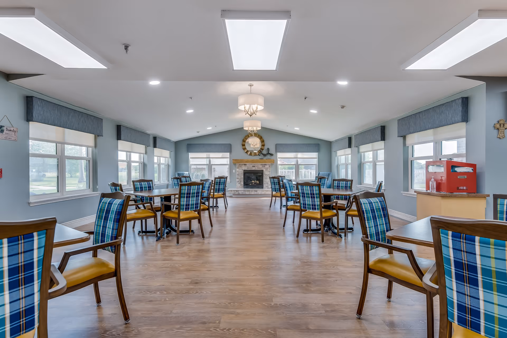 A spacious dining room with multiple wooden tables and chairs featuring blue and green plaid upholstery. The room has large windows with blue valances allowing natural light to fill the space. At the far end, there is a stone fireplace with a decorative round mirror above it. The ceiling has recessed lighting and two hanging light fixtures. The floor is wooden, and there is a red beverage dispenser on a counter to the right.