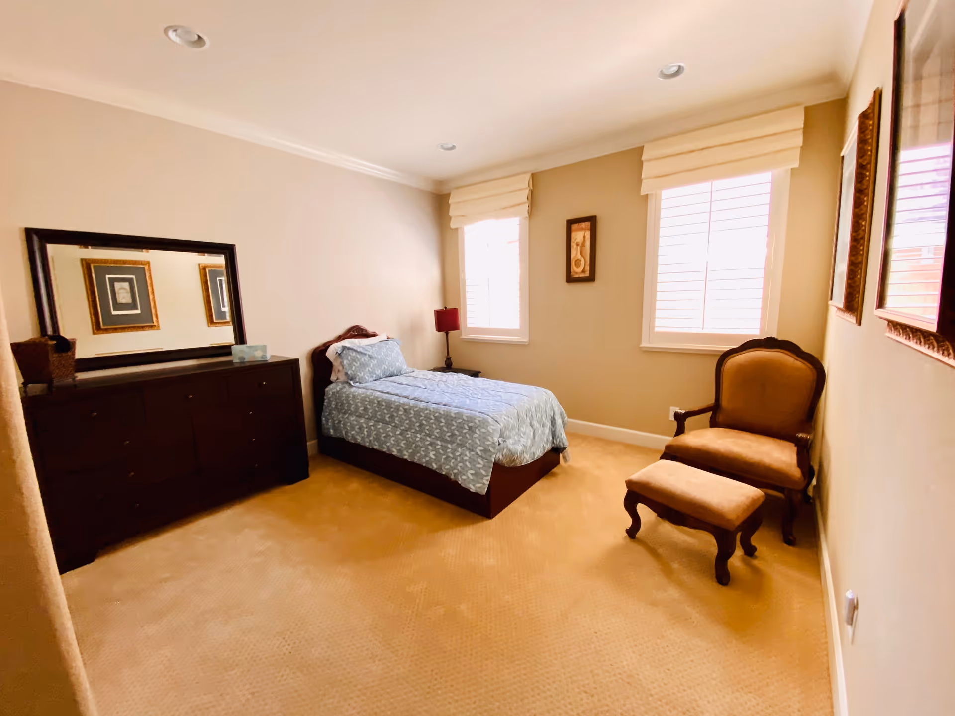 Sunlit bedroom with a single bed, a dark dresser with mirror, and an upholstered chair with ottoman.