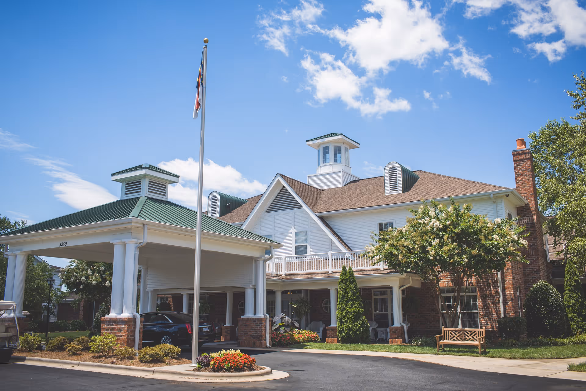 Exterior view of a senior living facility named Homestead Hills on a sunny day with a blue sky and some clouds. The building features a covered entrance with white columns and a green roof, a flagpole with a flag, brick and white siding, and landscaped greenery including bushes, flowers, and a tree with a wooden bench underneath.