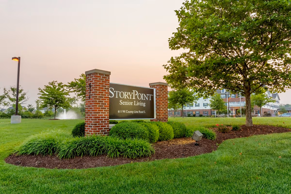 Outdoor view of StoryPoint Senior Living facility sign with landscaped greenery, trees, and a building in the background during sunset.