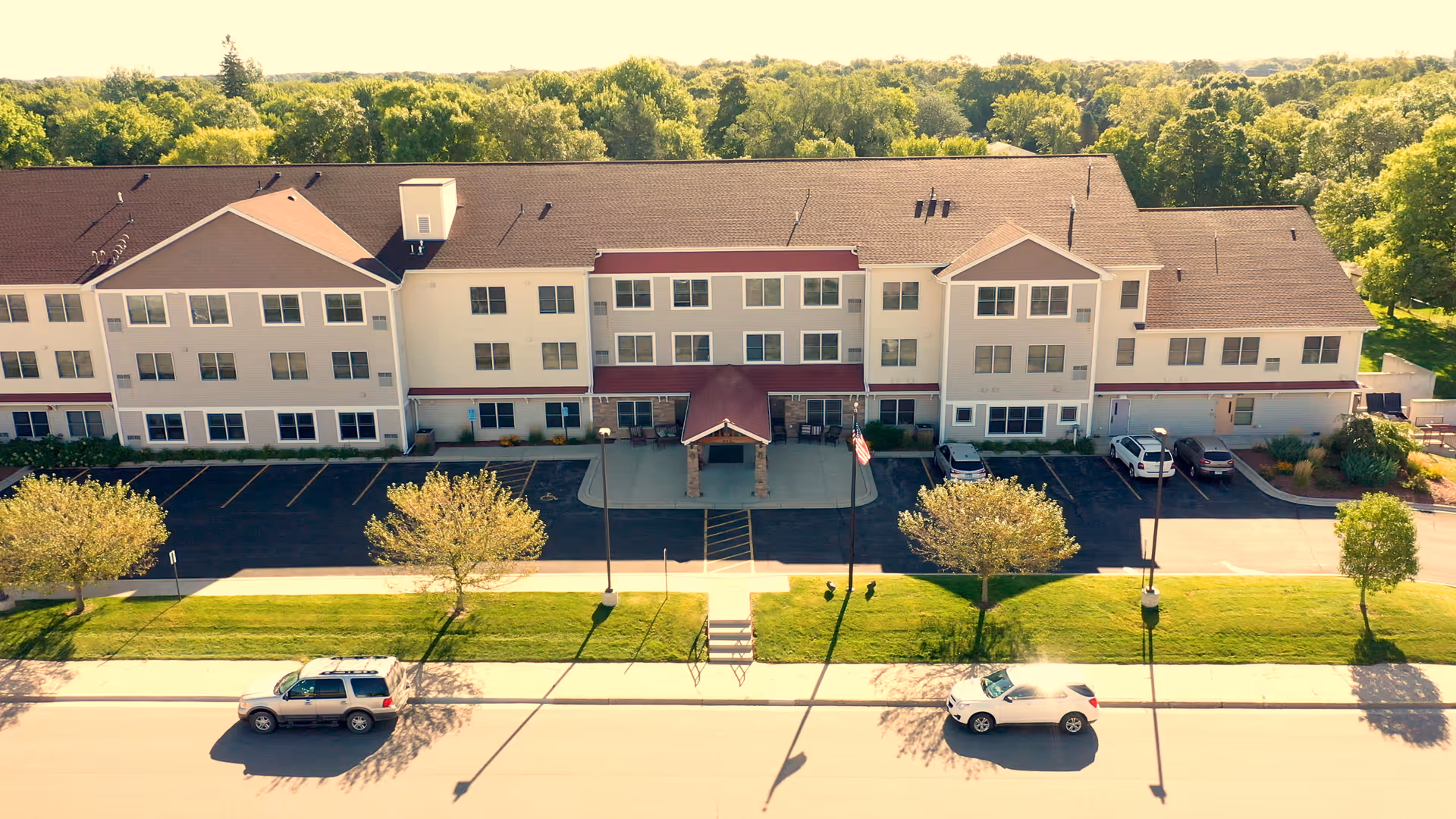 Front exterior view of a three-story senior living facility building with a covered entrance, parking spaces, and a well-maintained lawn with trees and street lamps. Two cars are parked on the street in front of the building, and a dense forest is visible in the background.