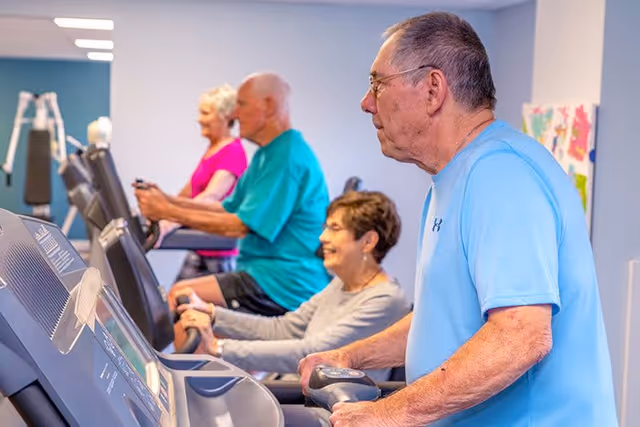 Four elderly individuals exercising on treadmills in a fitness room, with a light blue wall and exercise equipment in the background.