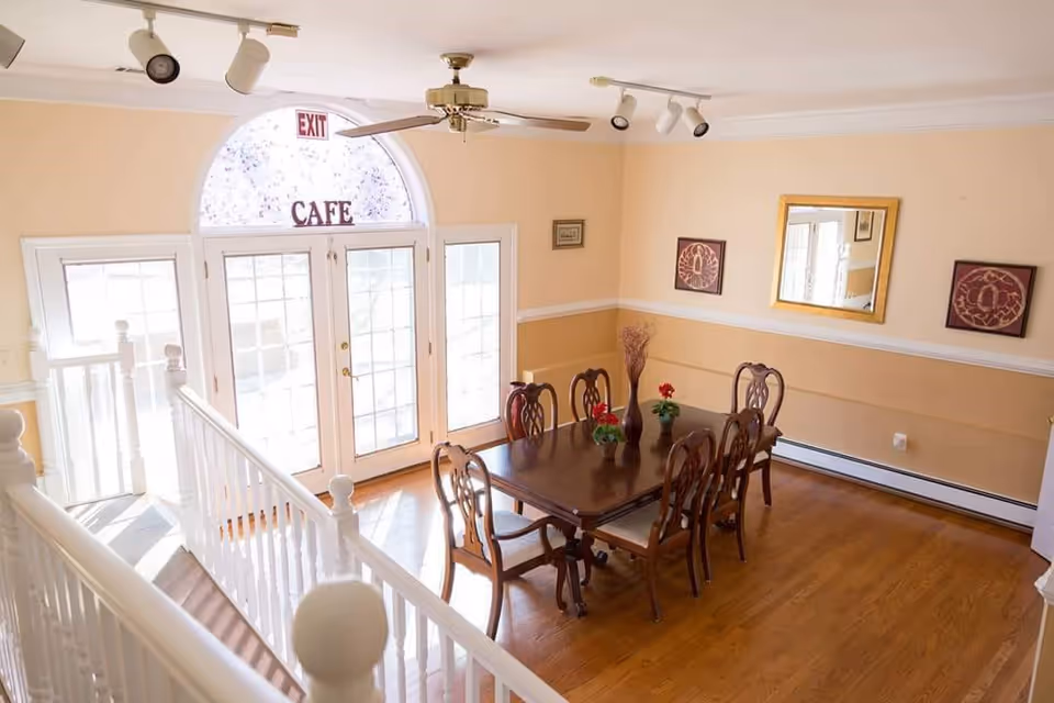 A bright dining room with a wooden table and six matching chairs. The table is decorated with small flower pots and a vase with dried plants. There is a large windowed door with an arched window above it, labeled 'CAFE' and an exit sign. The walls are painted beige with white trim, and there are framed artworks and a mirror on the walls. A white railing and staircase are visible in the foreground.