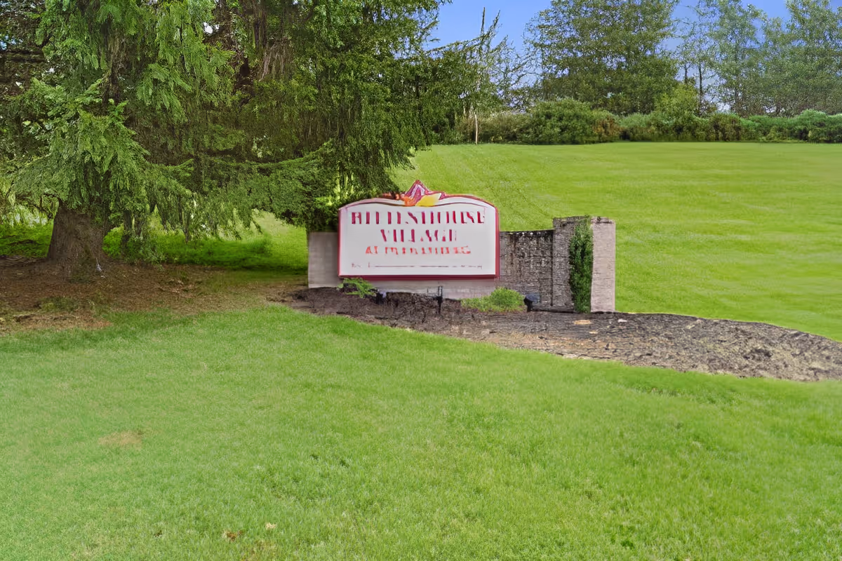Outdoor view of a green grassy area with a large tree on the left and a stone sign that reads 'Rittenhouse Village At Muhlenberg' surrounded by shrubs and landscaping.