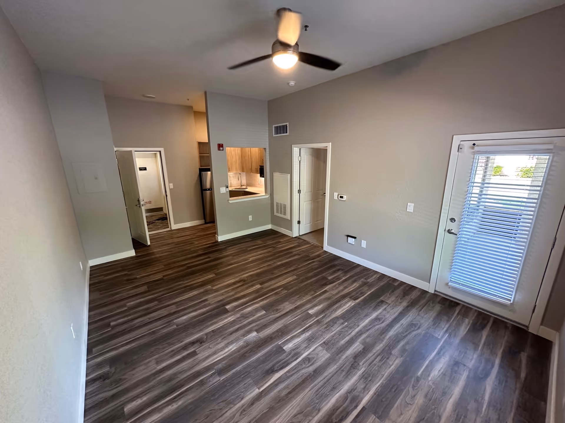 Empty room with wood-patterned flooring, beige walls, a ceiling fan with light, a small kitchen area with a refrigerator and cabinets, an open door leading to another room, and a glass door with blinds leading outside.