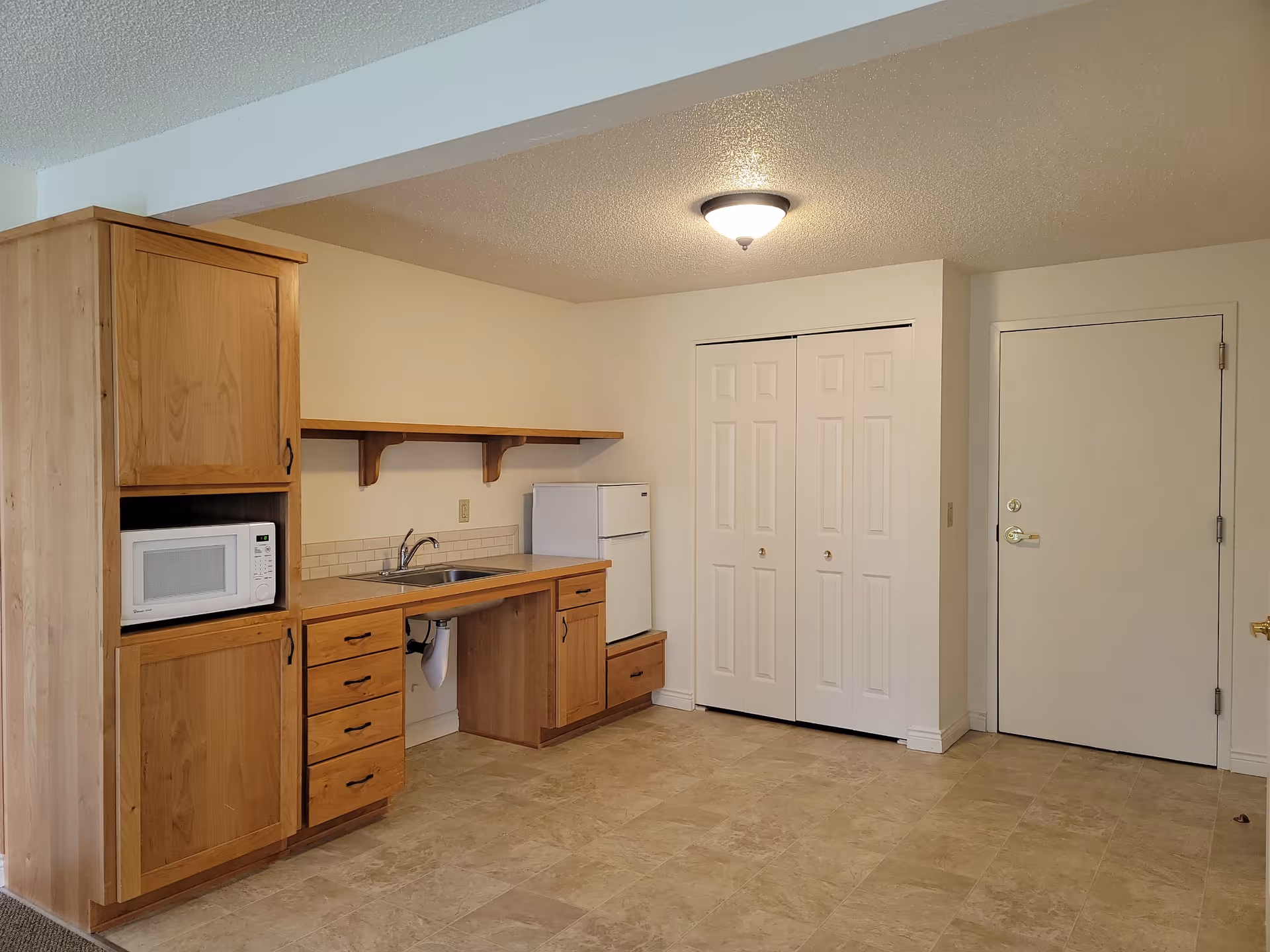 Small kitchenette area with wooden cabinets, a sink, microwave, mini-fridge, closet doors and an entry door in a studio-style room.
