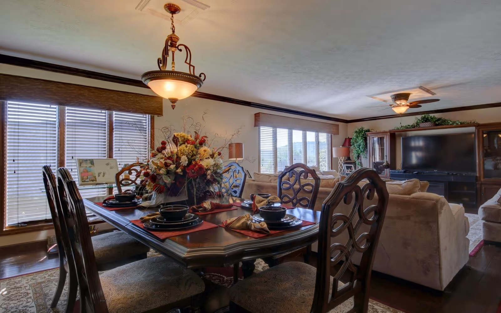 A cozy living and dining room area with a wooden dining table set for six, featuring dark wood chairs with intricate backs and a floral centerpiece. The room has large windows with blinds, beige sofas, a large flat-screen TV, and decorative plants and lamps.