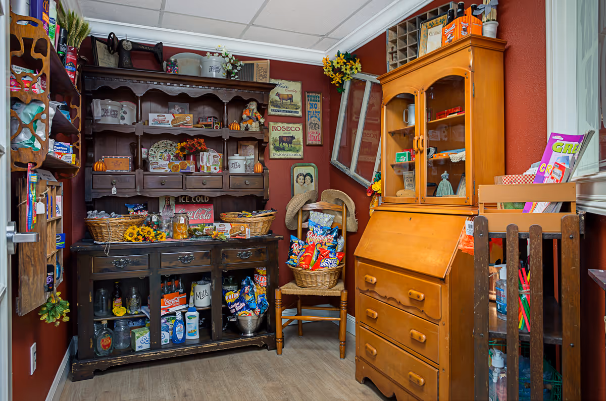 A cozy room with vintage wooden furniture including a dark wood hutch filled with decorative items and snacks, a wooden chair holding baskets of chips, and a light wood cabinet with glass doors displaying various items. The walls are painted a warm red and decorated with rustic signs and a tilted window frame. The floor is light-colored wood.