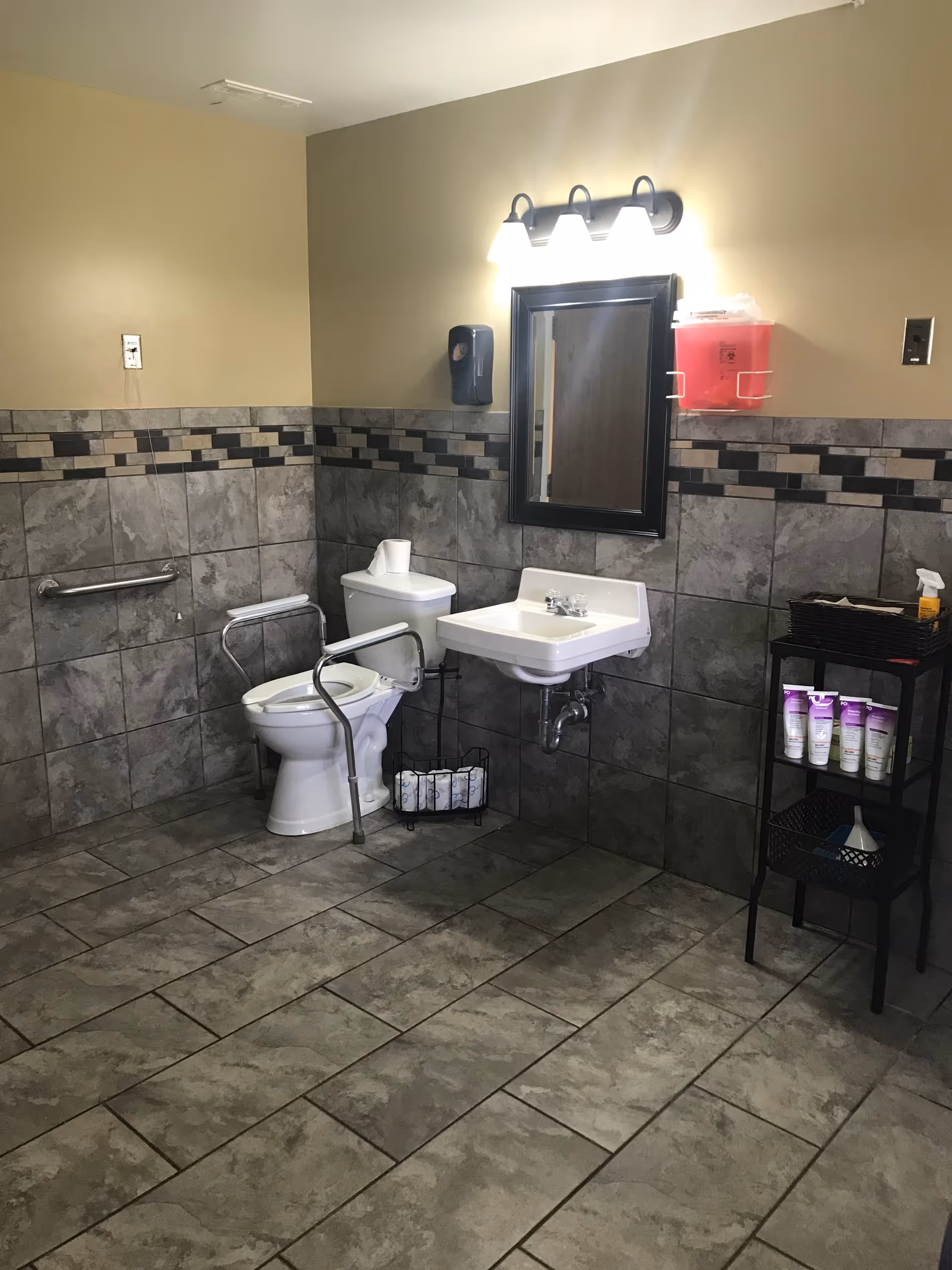 Accessible tiled bathroom with a toilet and safety rails, wall-mounted sink and mirror, and a small shelving unit with toiletries.