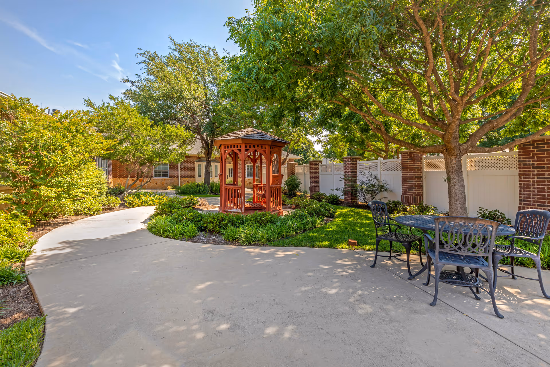 Outdoor garden area at Oak Ridge Alzheimer's Special Care Center featuring a curved concrete pathway, a red wooden gazebo surrounded by greenery, a large tree providing shade, and a black metal table with four chairs on a patio.