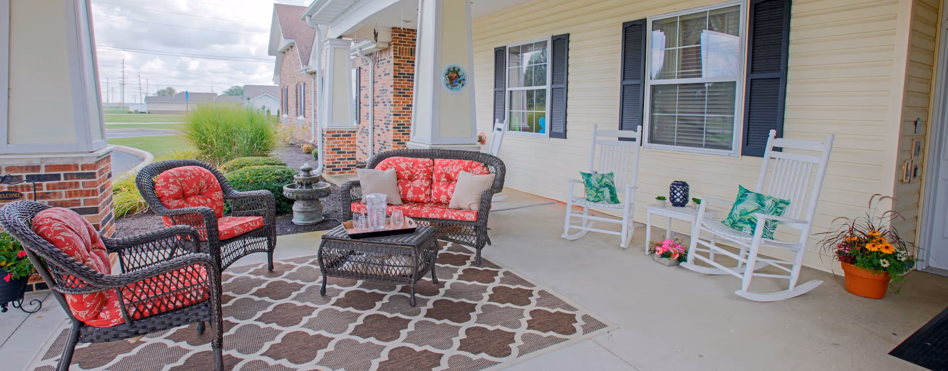 Covered front porch with wicker seating and red cushions, white rocking chairs, potted plants and a patterned rug.