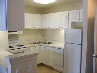A compact kitchen with white cabinetry, white appliances including a refrigerator, stove, and dishwasher, a double sink, and beige countertops and walls.