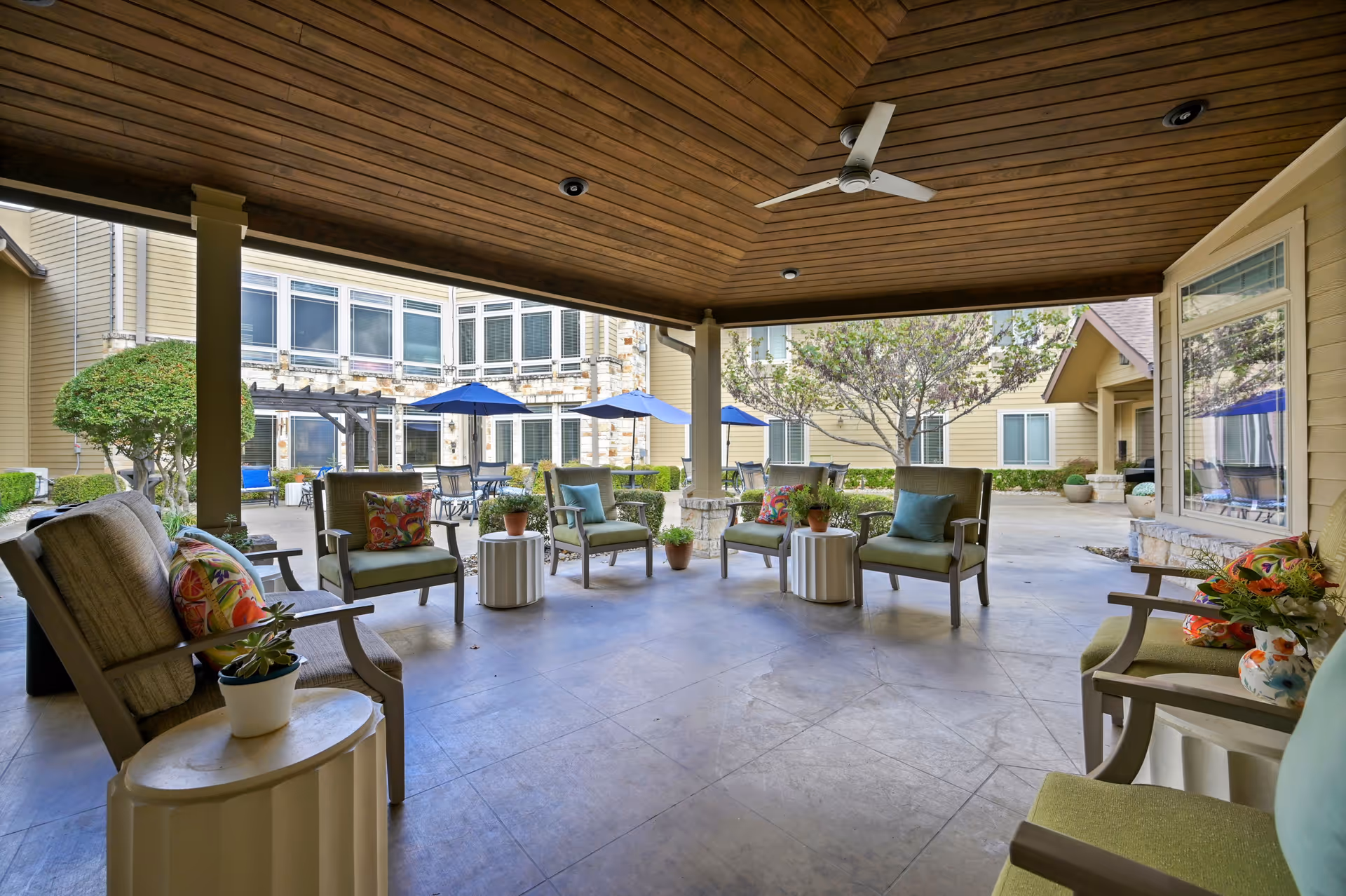 Covered outdoor seating area with cushioned chairs and small tables, overlooking a courtyard with patio tables, blue umbrellas, trees, and a building with large windows.