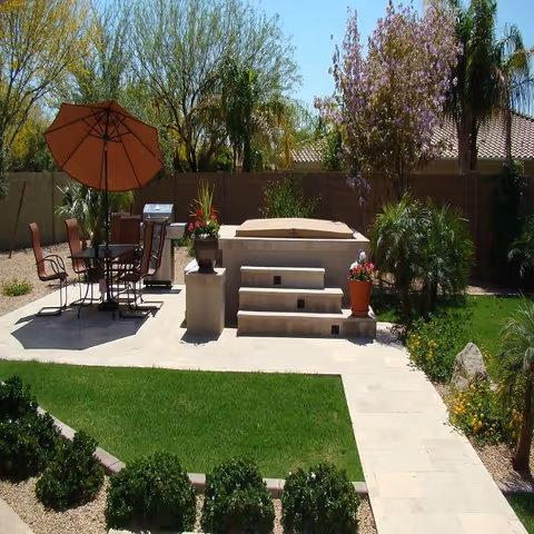 Outdoor patio area with a round table and four chairs under a large brown umbrella, a barbecue grill, steps leading up to a raised platform, surrounded by green grass, bushes, palm trees, and flowering plants under a clear blue sky.