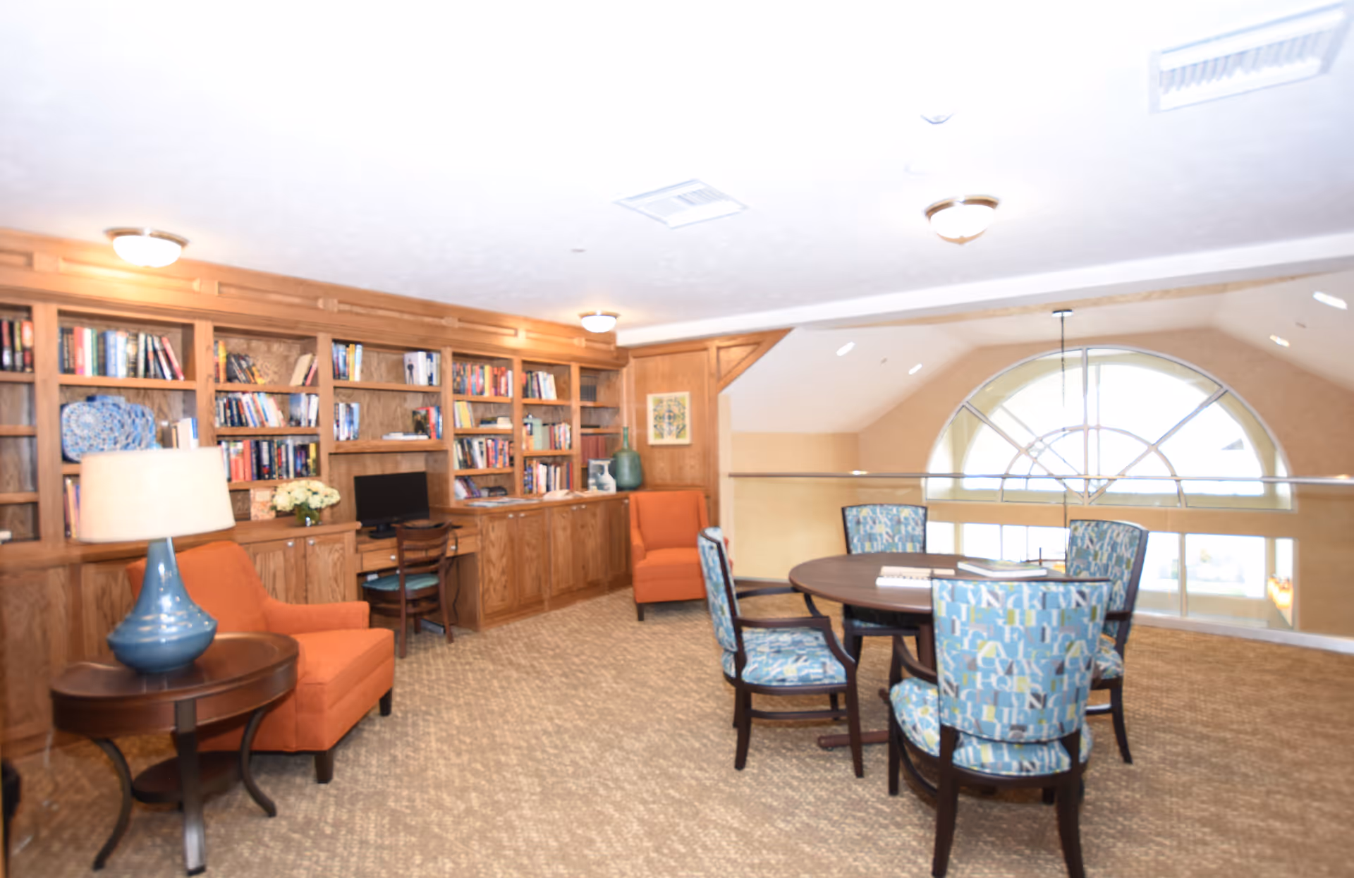 A cozy interior room with a large wooden bookshelf filled with books and decorative items along one wall. There are two orange armchairs, a small round table with a blue lamp, and a wooden desk with a computer. In the center of the room is a round table surrounded by four patterned chairs. The room has beige carpet and a large arched window letting in natural light.