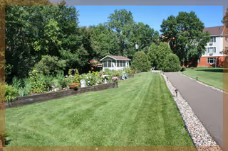 A well-maintained outdoor garden area with a green lawn, flower beds, and a small white gazebo. There is a paved walking path bordered by rocks on one side, and trees and shrubs surround the garden. Part of a multi-story building is visible in the background.