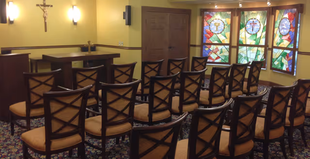 Interior view of a small chapel or prayer room with rows of wooden chairs facing an altar. The room has yellow walls, a crucifix on the wall, wall-mounted lights, and stained glass windows with religious symbols.