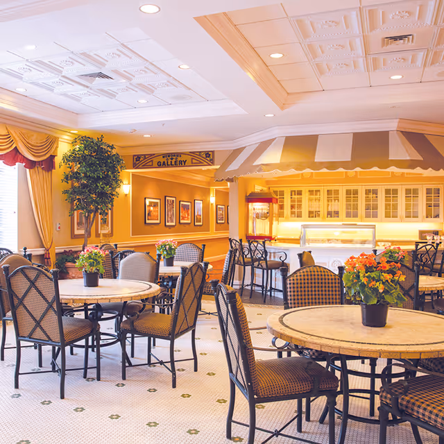 A bright and inviting dining area with round tables and cushioned chairs, each table adorned with a small potted plant with colorful flowers. The room features a decorative ceiling, warm yellow walls with framed pictures, and a counter area with a striped awning and a popcorn machine. Large windows with draped curtains allow natural light to fill the space.