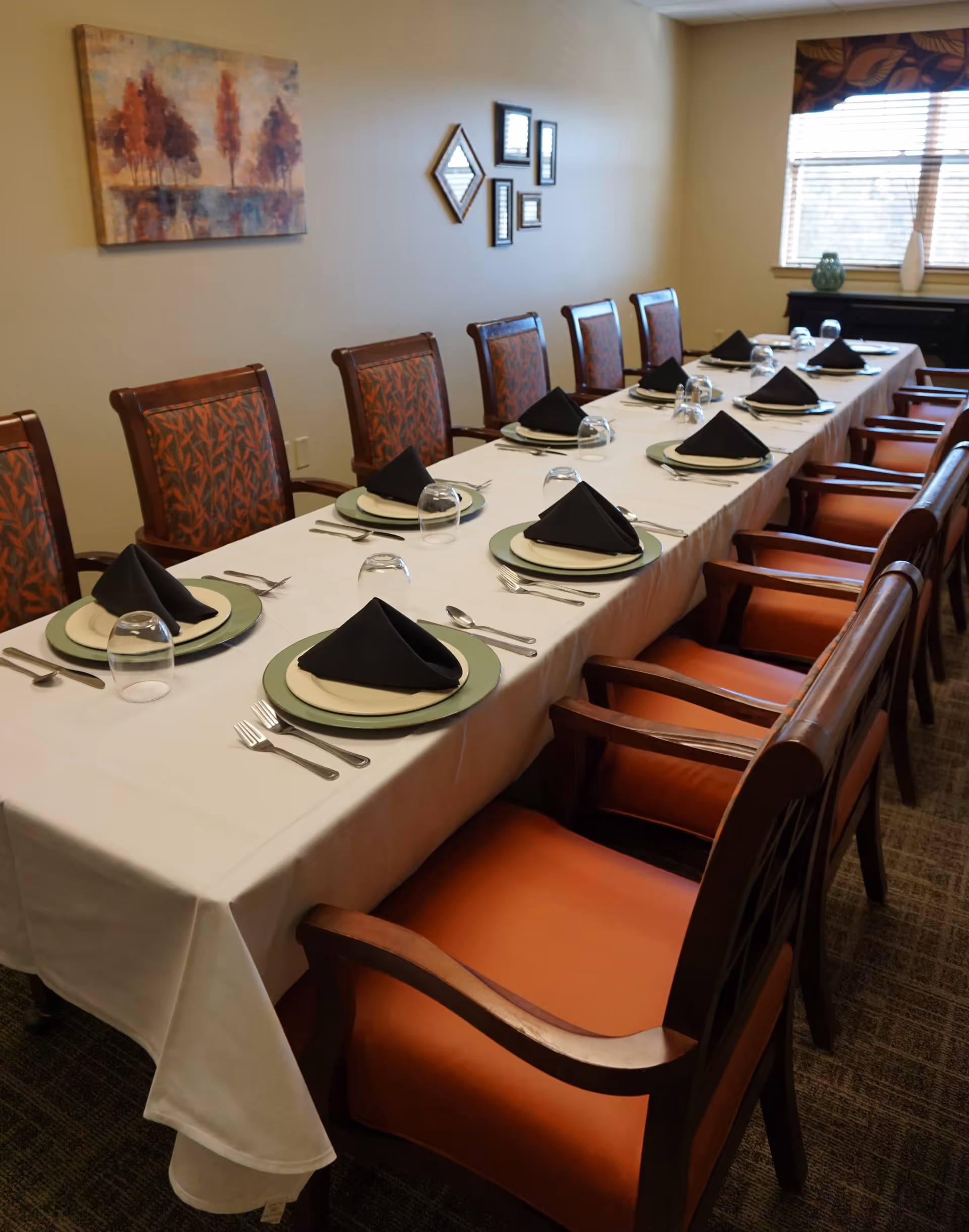 Long dining table set for a meal with plates, inverted glasses and black folded napkins, surrounded by upholstered wooden chairs in a dining room.