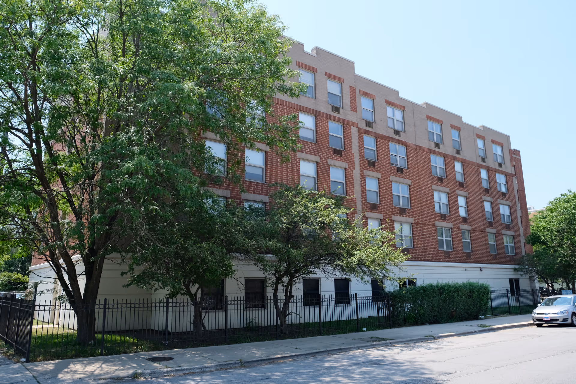 Exterior view of a five-story red-brick apartment building with trees, a fence, and a parked car on a sunny street.