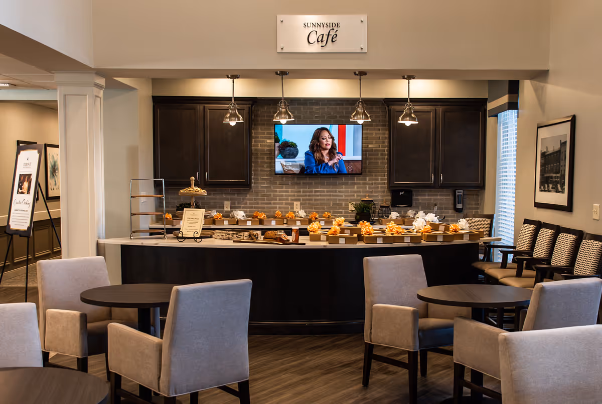 Interior view of a senior living facility café named Sunnyside Café featuring a counter with gift boxes decorated with bows, a mounted TV screen showing a woman speaking, dark wood cabinets, pendant lights, round tables with beige upholstered chairs, and a seating area with chairs along the wall.